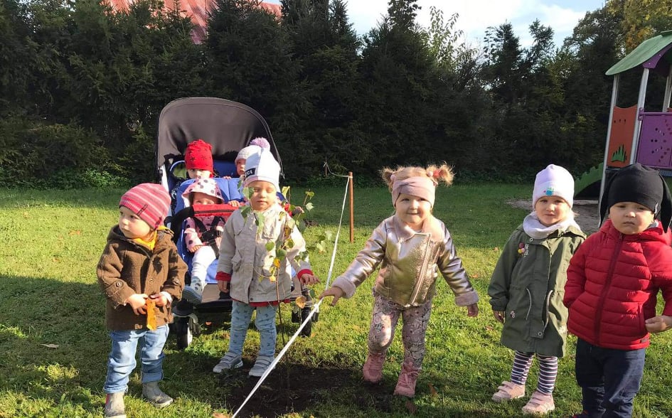 Dzieci z grupy Krasnali i Muchomorków stoją w pobliżu nowo posadzonego drzewka z okazji Dnia Drzewa. 