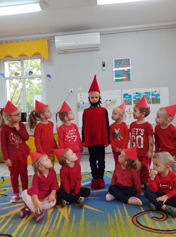 dzieci w wieku 3,4 lata ubrane na czerwono, z czerwonymi czapeczkami na głowie pozują do zdjęcia z dzieckiem przebranym  za Krasnala Hałabałę