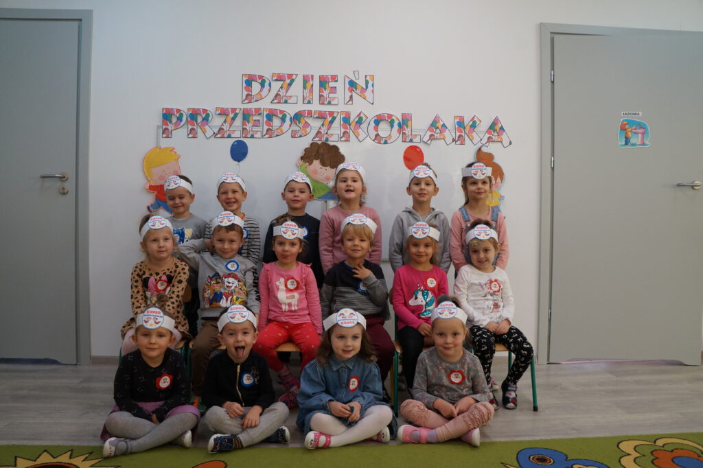 dzieci z opaskami na głowie z napisem super przedszkolak ustawione na tle tablicy z napisem dzień przedszkolaka