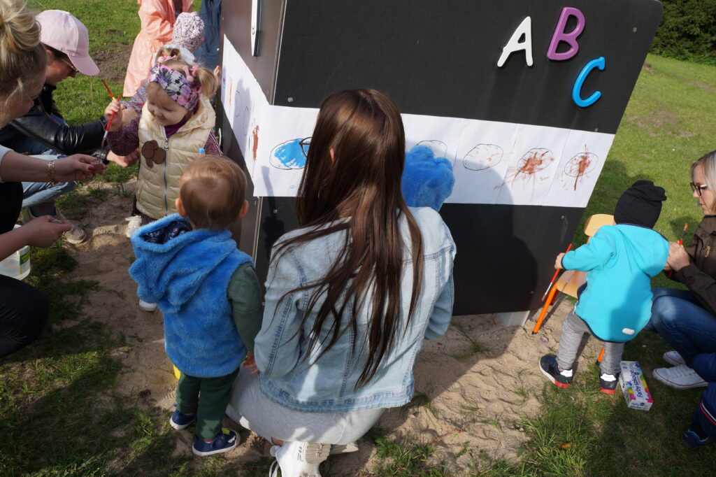 Dzieci wraz z rodzicami malują farbami na tablicy w ogrodzie ilustrację ziemniaka.
