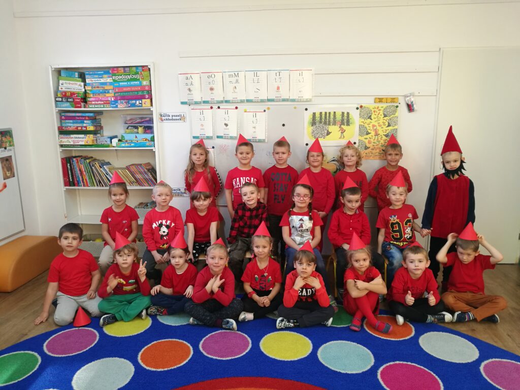 dzieci w wieku 5,6 lat ubrane na czerwono, z czerwonymi czapeczkami na głowie pozują do zdjęcia z dzieckiem przebranym  za Krasnala Hałabałę