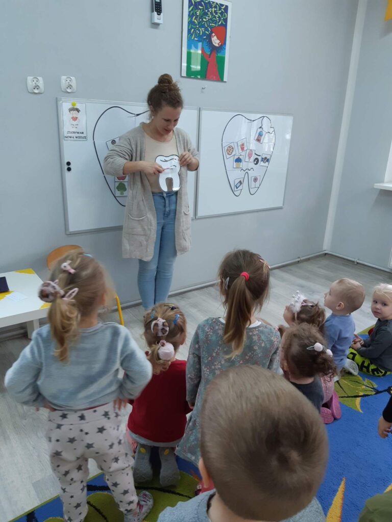 Nauczycielka pokazuje grupie dzieci jak prawidłowo myć zęby.