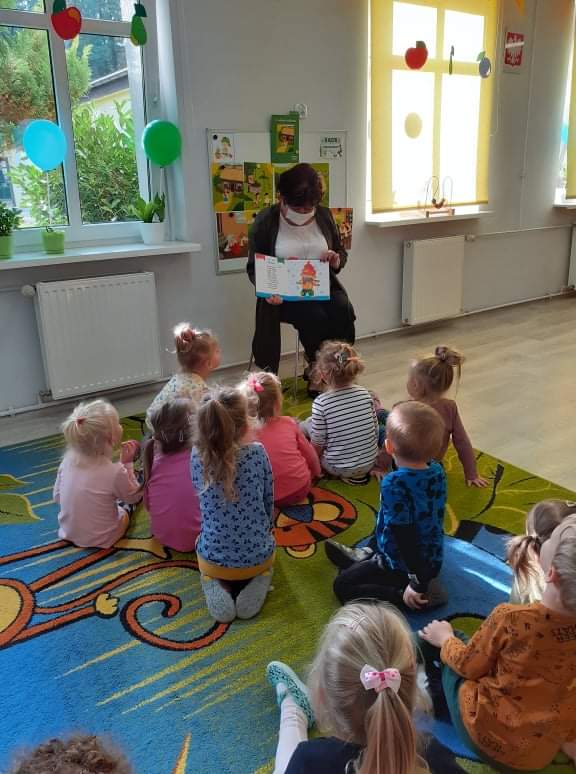 kobieta pokazuje dzieciom książkę, dzieci w wieku przedszkolnym zwrócone w stronę kobiety
