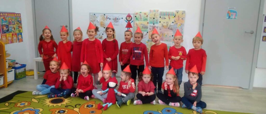 dzieci ustawione na tle tablicy z kolorowymi obrazkami, dzieci ubrane na czerwono, z czerwonymi czapeczkami na głowie, pozują DO ZDJĘCIA Z MASKOTKĄ KRASNALA HAŁABAŁY