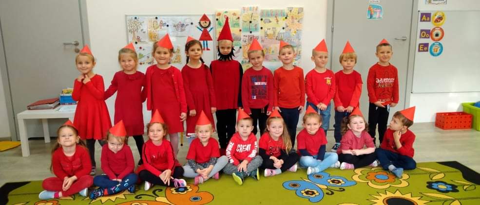 dzieci w wieku 4,5 lat ubrane na czerwono, z czerwonymi czapeczkami na głowie pozują do zdjęcia z dzieckiem przebranym  za Krasnala Hałabałę