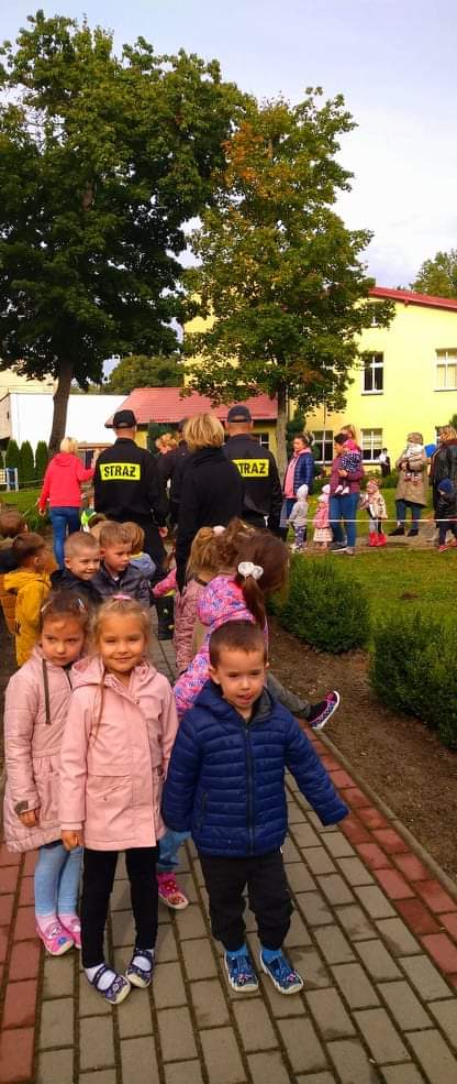 grupa dzieci, dorosłych i strażacy stoją na chodniku przed przedszkolem 