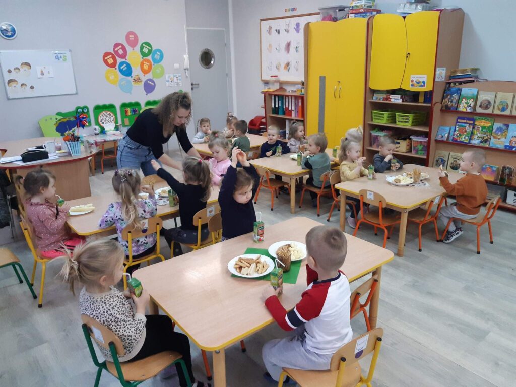 Dzieci siedząc przy stolikach spożywają słodki poczęstunek.