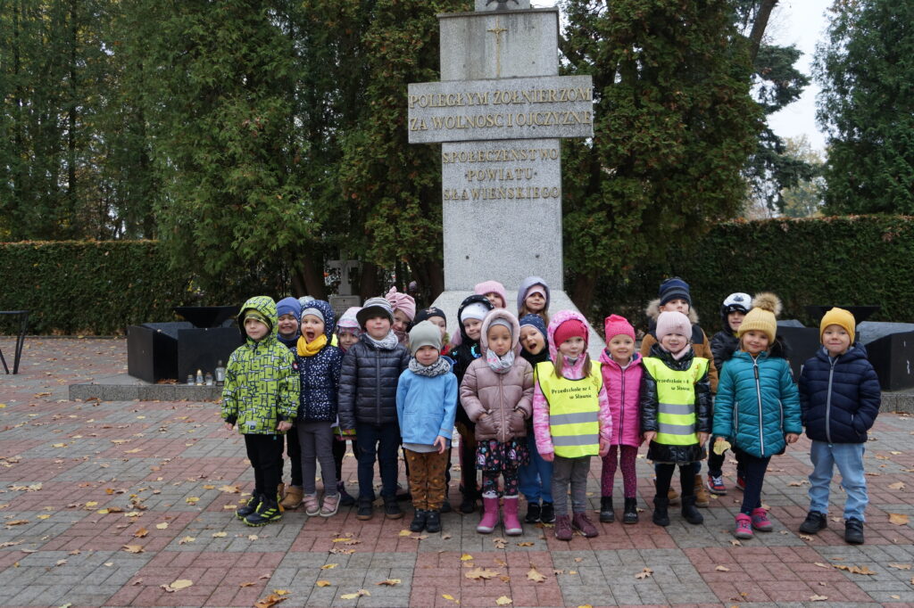 Grupa dzieci stoi przed pomnikiem żołnierskim na cmentarzu