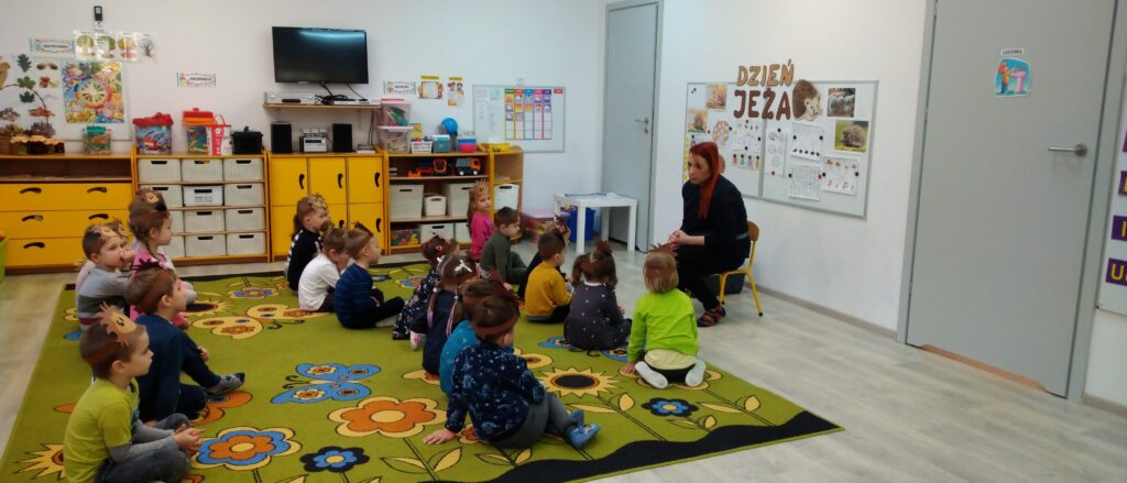 Dzieci siedzą w przedszkolu , w rzędach na dywanie i rozmawiają z Panią-opiekunem małego jeża