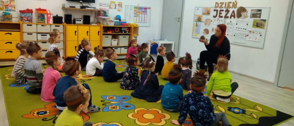 Pani opiekunka jeża trzyma na rękach małego jeżyka, przedszkolaki siedzące na dywanie słuchają ciekawostek na jego temat 
