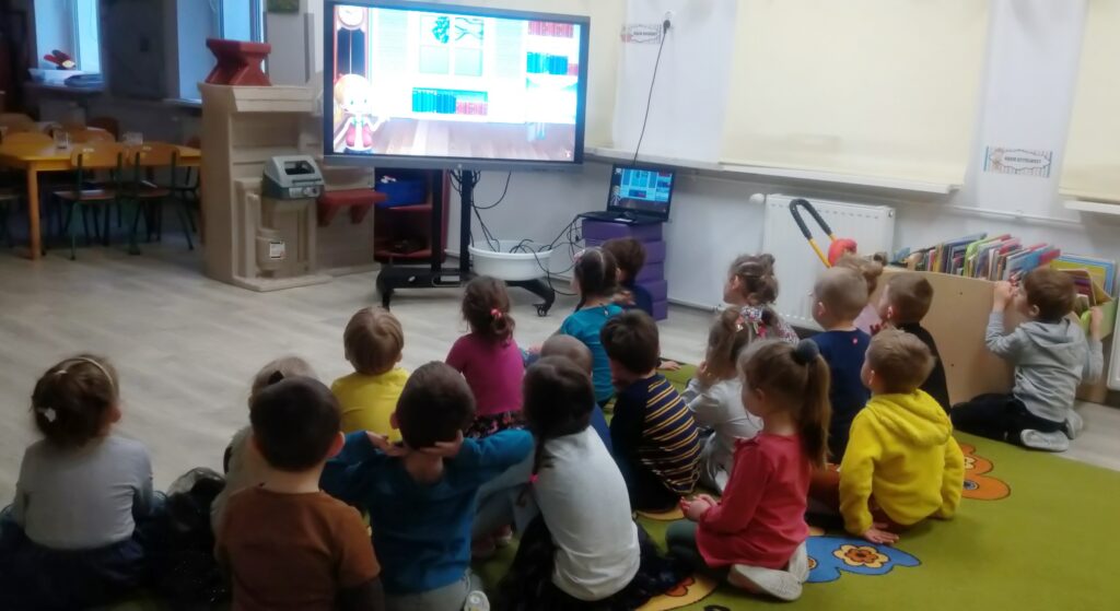 Dzieci siedzą na dywanie i oglądają na tablicy multimedialnej prezentację o andrzejkach