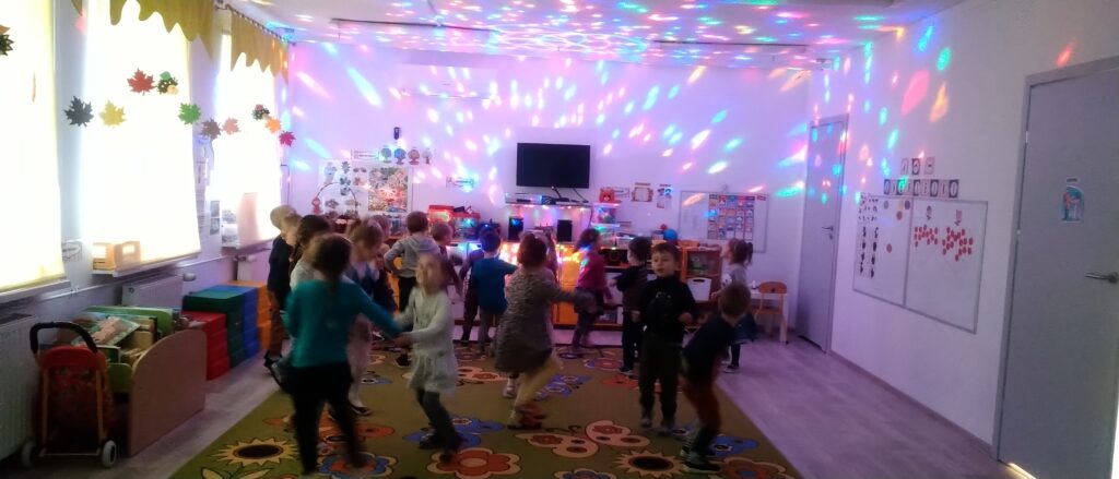 Dzieci tańczą w swojej sali przedszkolnej , trzymają się za ręce lub podskakują w rytm muzyki, na ścianach i suficie widać kolorowe światełka