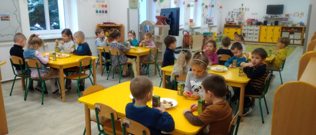 Dzieci siedzą przy stolikach, zjadają słodycze i  piją soczek - poczęstunek andrzejkowy