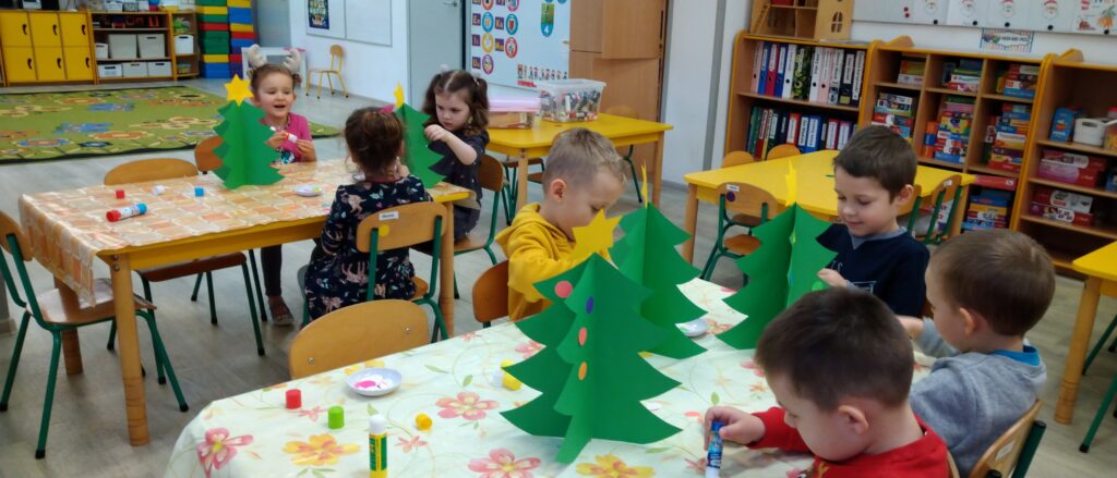 Dzieci siedzą przy stolikach , wykonuja świąteczną ozdobę na stoły, naklejaja na papierową choinkę kolorowe bombki