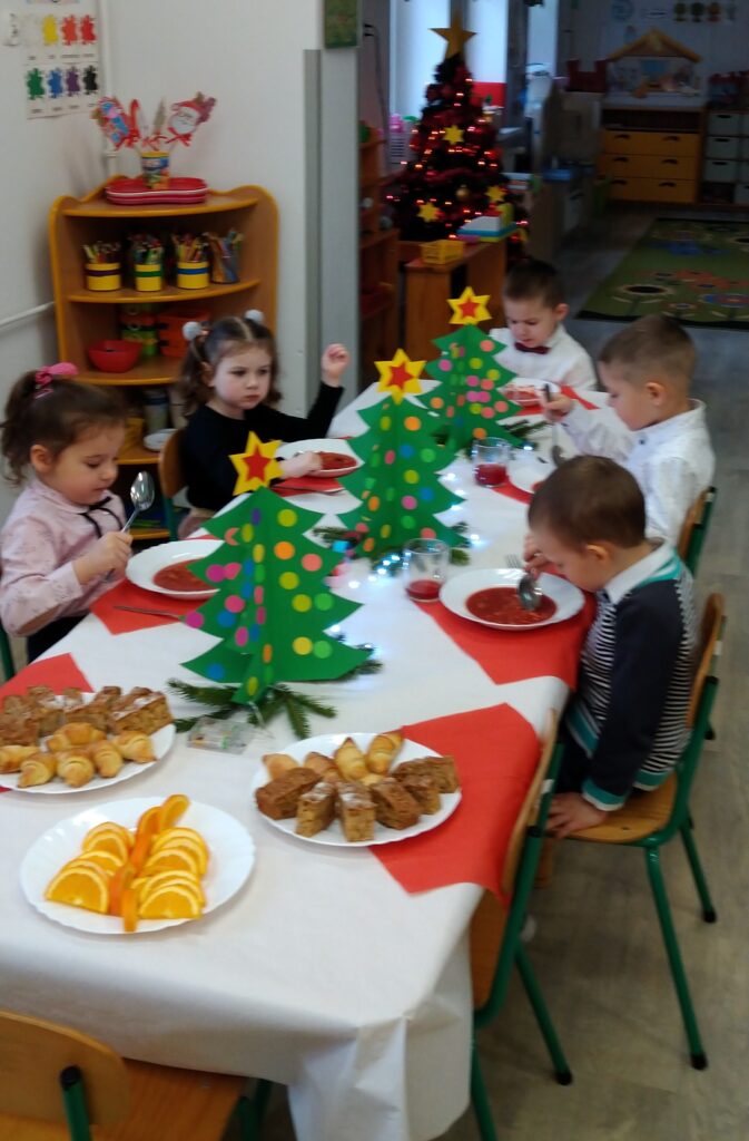Dzieci siedzą pzry stole jedzą świąteczny obiad. Stół odświętnie nakryty, dzieci sa ubrane w stroje galowe