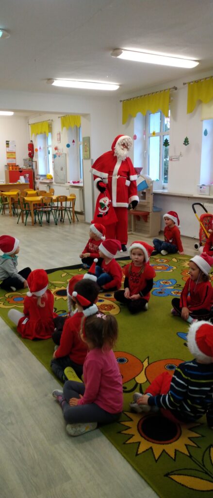 Dzieci siedzą na dywanie, witaja gościa- Św. Mikołaja, który wszedł do sali pzredszkolnej
