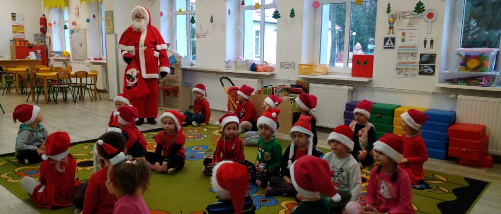 Dzieci siedzą w rzędach na dywanie, witaja Mikołaja, który ich odwiedził.
