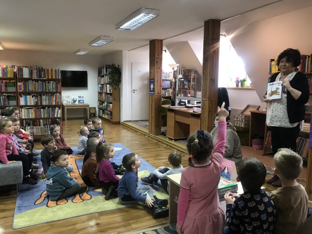 Dzieci z pzredszkola poszły na wycieczkę do biblioteki. Siedzą na dywanie w sali przeznaczonej właśnie dla najmłodszych. Pani bibliotekarka prezentuje im wybraną przez siebie książkę i opowiada, jak zostac czytelnikiem.