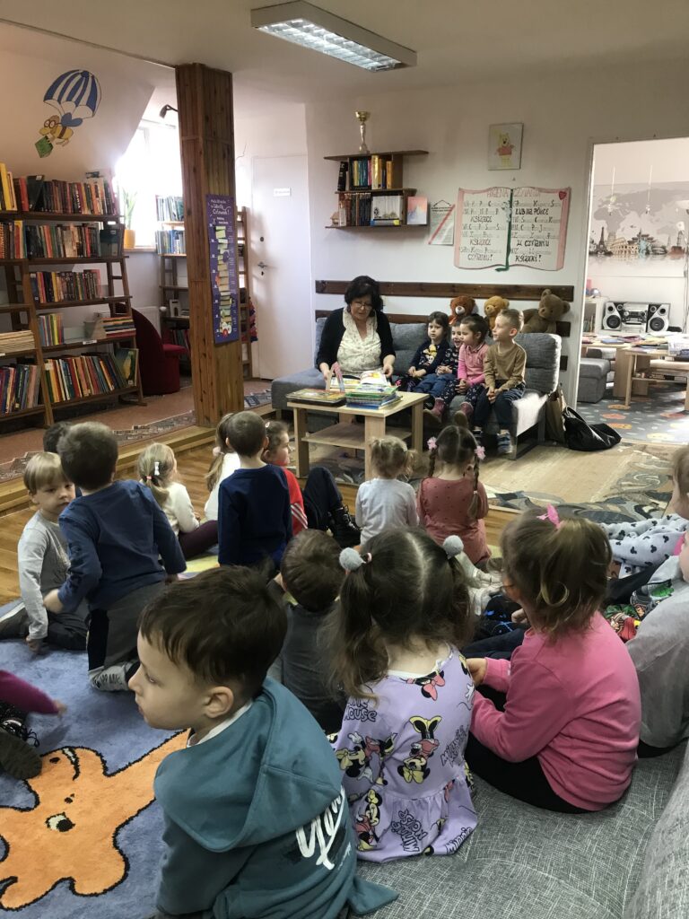Dzieci zwiedzaja biblioteke miejską. Siedza w sali bibliotecznej na dywanie i na kanapach. Obok siedzi Pani pracująca w bibliotece i czyta im wiersze. 