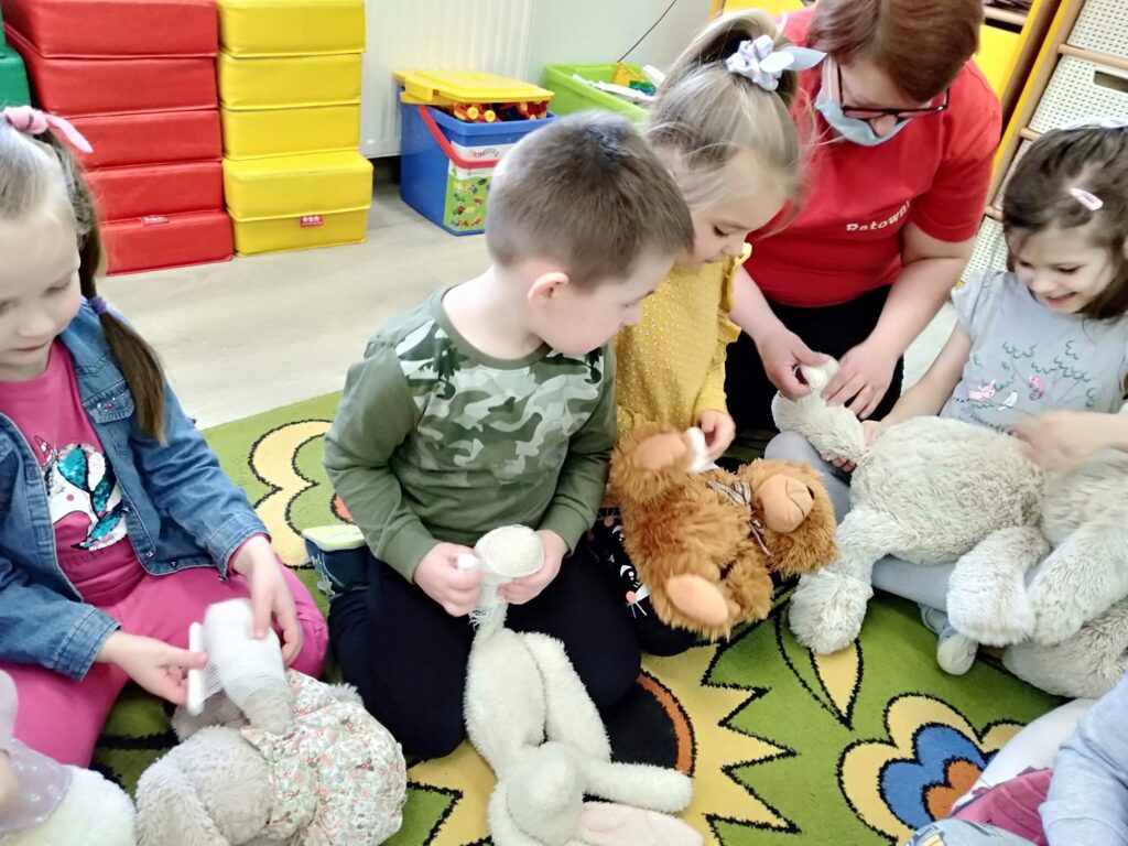 Dzieci siedzą na dywanie,  trzymają przed soba pluszaka. Kazdy przedszkolak próbuje zabandażowac mu chorą łapkę. Zaproszona Pani ratownik pomaga w tych czynnościach