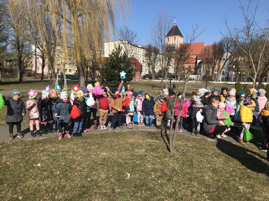 Dzieci z przedszkola spacerują w parku, trzymając w rękach kolorowe pompony i kwiaty. Witaja Panią Wiosne.