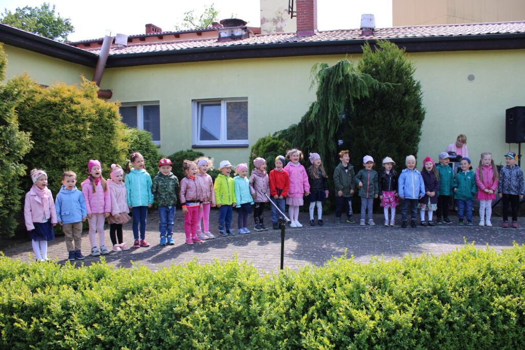 Zdjęcie przedstawia uroczystość z okazji Dnia Mamy i Taty w przedszkolnym ogrodzie. Dzieci z grupy II stoją w rzędzie na cenie mini-amfiteatru i śpiewaja piosenkę dla swoich rodziców oraz składają życzenia mamie i tacie. 