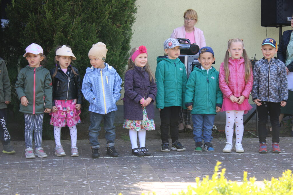 Dzieci ( 8 osób) stoją na scenie przedszkolnego amfiteatru w ogrodzie i recytują wiersz dla swoich rodziców z okazji Dnia Mamy i Taty. 