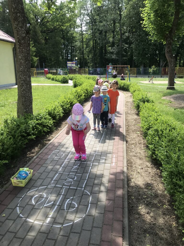 Dzieci bawią się w ogordzie przedszkolnym w czasie pięknej pogody. Uczą się skakać po narysowanym na chodniku " chłopku", albo innymi słowy - "grają w klasy:", Stoją w rzędzie i kolejno obunóż przeskakuja po określonyhc polach narysowanych kredą na chodniku. 