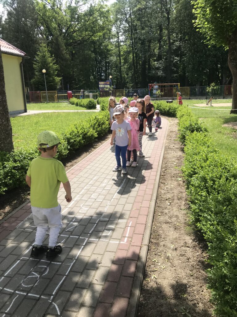 Zdjęcie przedstawia dzieci w ogrodzie przedszkolnym, skaczących po chodniku i bawiących się w " klasy" Przedszkolaki kolejno ustawione w rzędzie przeskakują po polach oznaczonych cyfrą. 