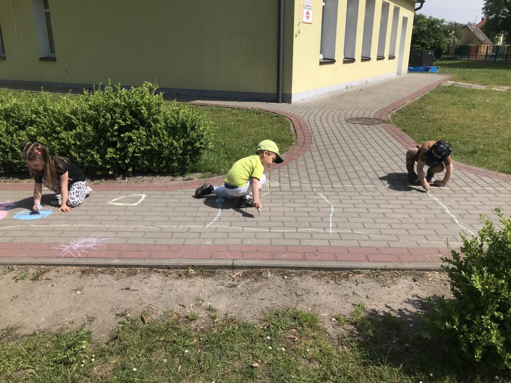 Troje dzieci rysuje kolorowa kredą na chodniku przed budynkiem przedszkola. Dzieci bawią się w czasie zabaw w ogrodzie rekreacyjnym.