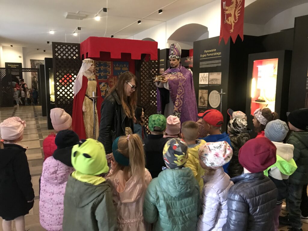 Dzieci z przedszkola słuchają opowieści pani przewodnik o koronacji na króla. Znajdują się w zamkowej sali i przyglądają się ustawionym postaciom króla i królowej. Wokół widac wiele gablot w których znajdują się królewskie rekwizyty.