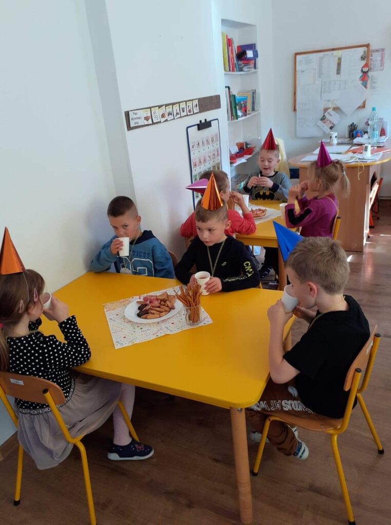 Dzieci w sali przedszkolnej siedzą przy stolikach zajadają słodki poczęstunek z okazji świeta Pzredszkolaka. Mają na głowach kolorowe czapeczki z okazji swojego swięta