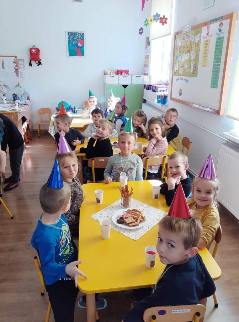 Dzieci siedzą przy stoliku w sali przedszkolnej, na głowach mają kolorowe czapeczki. Zjadaja przygotowany słodki poczęstunek z okazji święta przedszkolaka.