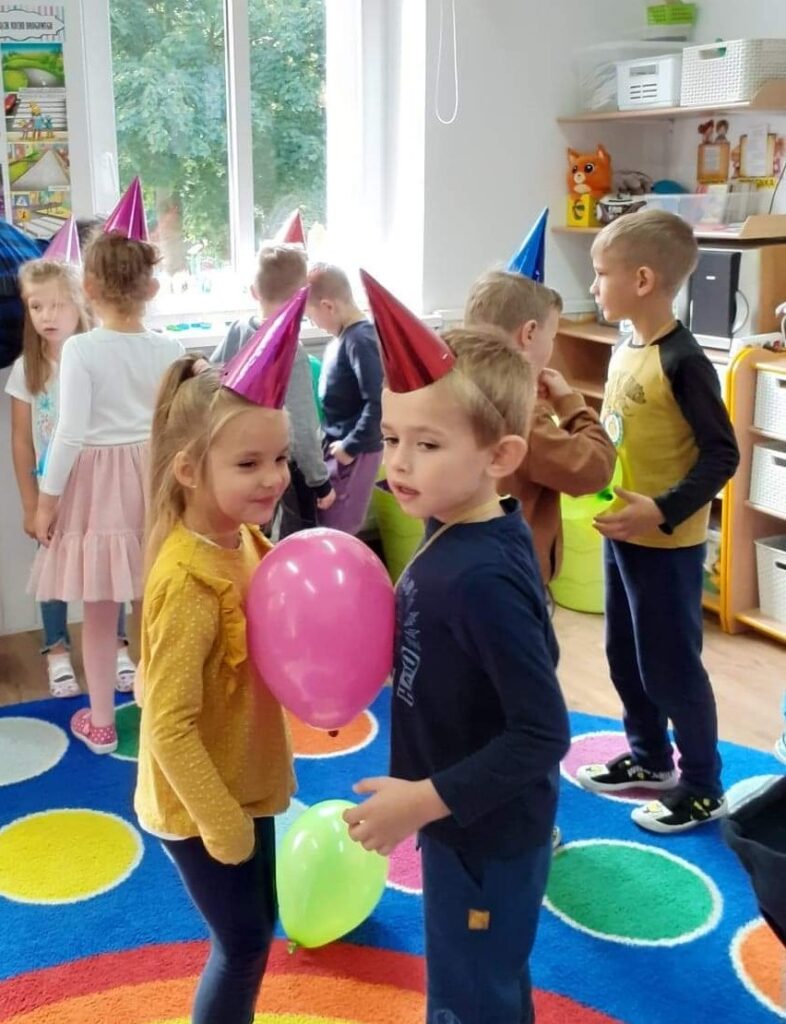 Dzieci w sali przedszkolnej uczestniczą w zabawie tanecznej z balonem. dzieci w kolorowych czapeczkach stoją w parach, brzuchami trzymaja balon. Zabawy zorganizowano z okazji dnia przedszkolaka. 