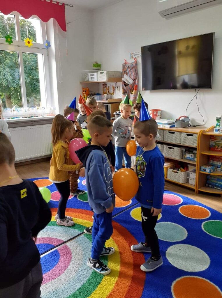 Dzieci obchodza w przedszkolu swoje święto. Tańczą na dywanie w parach trzymając brzuchem balonik. 