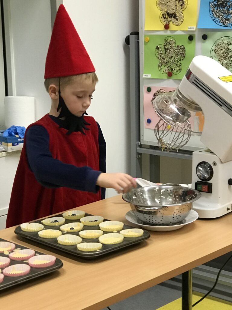 Chlopiec przebrany za Krasnala posypuje babeczki borówkami. Pomaga pani kucharce w przygotowaniu deseru