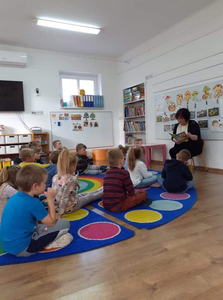 Dzieci w przedskolu siedzą na dywanie.  Odwiedziła ich pani bibliotekarka, ktora Z okazji Dnia głośnego Czytania czytała dzieciom w pzredszkolu wybrana bajkę , zachęcając ich do sięgania po ksiażki wspólnie z rodzicami