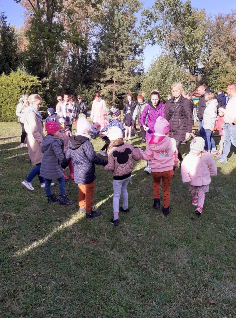 Dzieci wspólnie z panią i rodzicami bawwia się w przedszkolmnym ogrodzi z okazji swięta pieczonego ziemnaika. tańczą razem z rodzicami trzymając się za ręke.