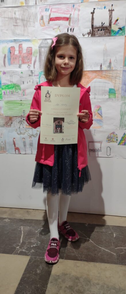 Dziewczynka z grupy starszaków trzyma w ręku dyplom , ktory zdobyła jako uczestnik konkursu plastycznego. Prezentuje otrzymaną nagrode na Zamku w Darlowie.