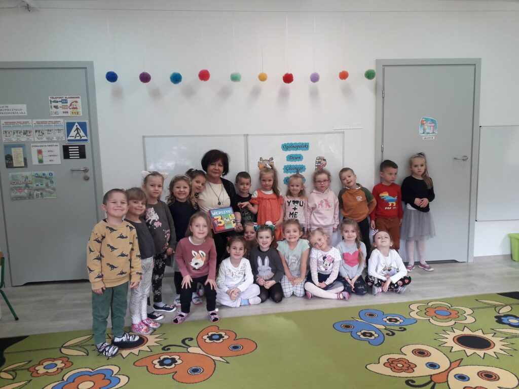 Grupa dzieci razem z panią Bogusią z Miejskiej Biblioteki w Sławnie na wspólnym zdjęciu w sali przedszkolnej.