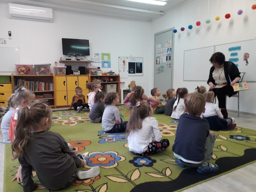 Pani bibliotekarka czyta dzieciom siedzącym na dywanie wybrane opowiadanie.