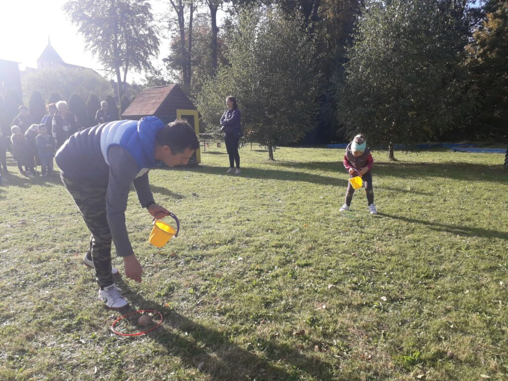 Tata i córka rywalizują ze sobą w konkurencji sadzenia ziemniaków.
W tle widać pozostałych uczestników zawodów sportowych.