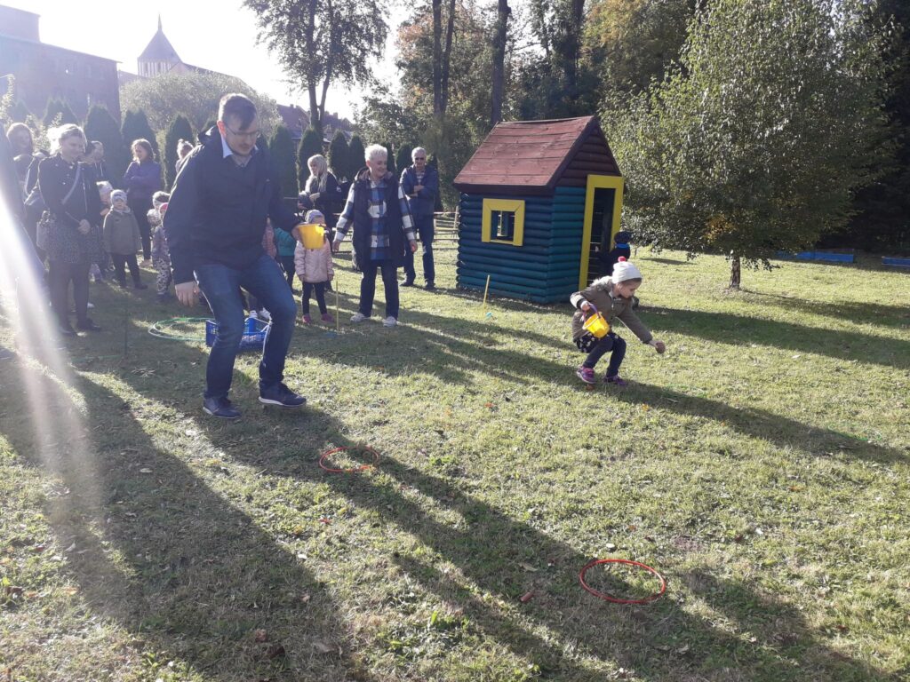 Tata rywalizuje z córką w konkurencji sadzenia ziemniaków. W tle pozostali uczestnicy zawodów sportowych.