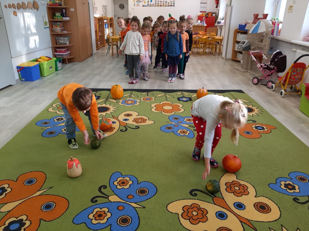 W sali przedszkolnej dwoje dzieci uczestniczy w zawodach sportowych polegających na toczeniu dyni slalomem. W tle cała grupa Średniaków oczekuje na swoją kolej kibicując kolegom.