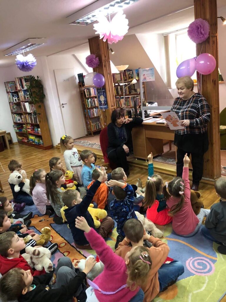 Grupa dzieci siedzi na dywanie w bibliotece miejskiej . Pzredszkolaki zostały zaproszone na obchody Święta MIsia. Panie bibliotekarki opowiadaja dzieciom ważne rzeczy o Misiach i ich pochodzeniu.