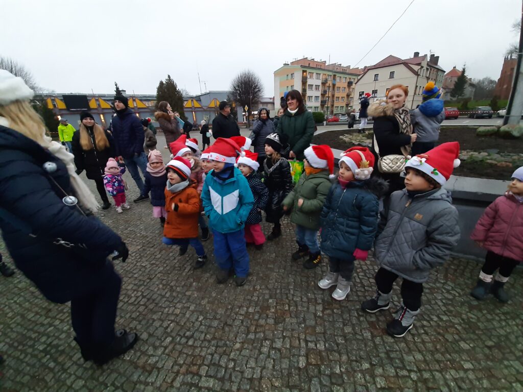 Grupa przedszkolaków, wśród innych osób, oczekuje na placu miejskim w Sławnie na uroczyste zapalenie lampek na choince miejskiej.