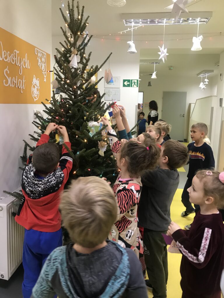 Dzieci zawieszają na choince wykonane przez siebie ozdoby. W tym roku zrobiły aniołki, ktore mają ozdabiać swiąteczne drzewko w holu przedszkola.