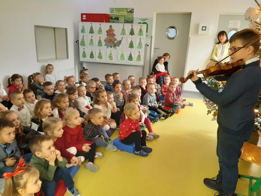 Dzieci siedzą w holu przedszkola słuchając koncertu kolęd.