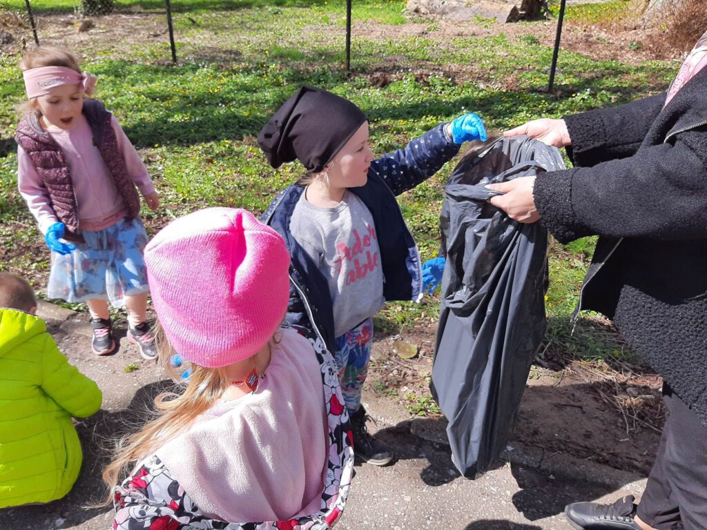 DZIECI W RAMACH AKCJI SPRZĄTAJĄ PARK W OKOLICY PRZEDSZKOLA WRZUCAJĄ ZNALEZIONE ŚMIECI DO WORKA