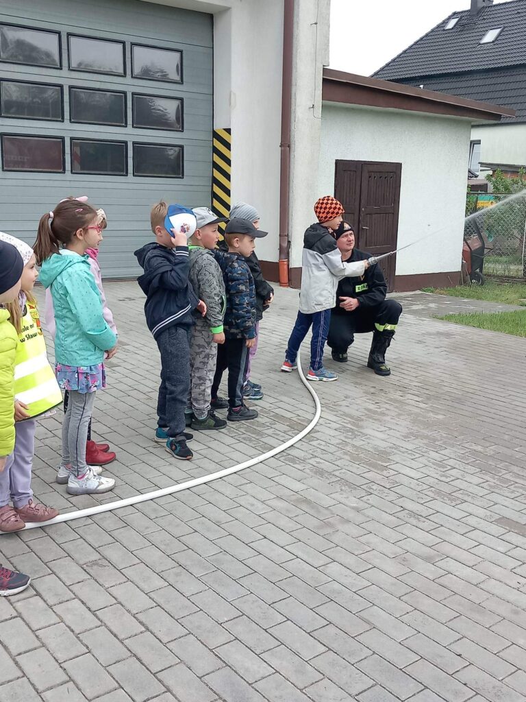 Dzieci na wycieczce u strazaków leja wężem strażackim wodę , próbja ugasic na niby pożar.